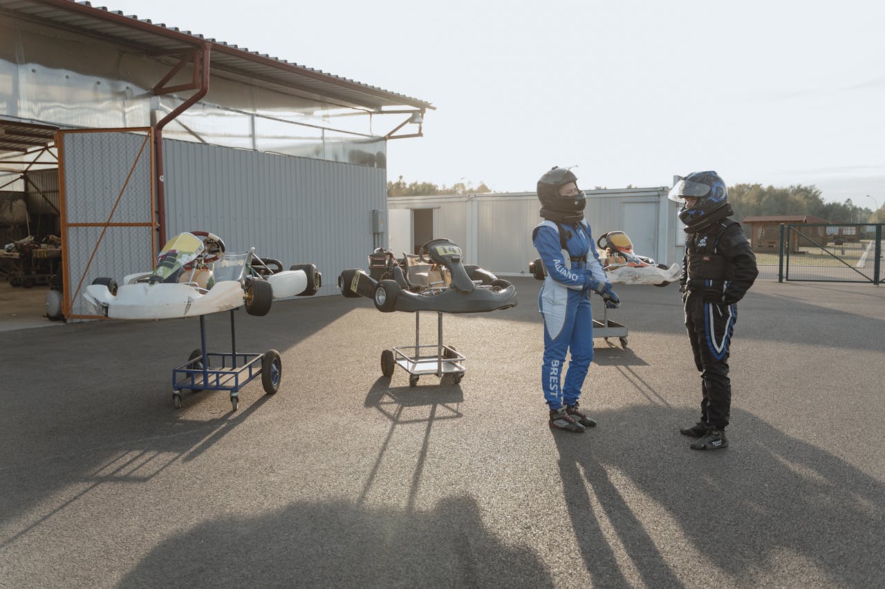 Two go-kart racers in protective gear standing trackside, preparing for a race outdoors.